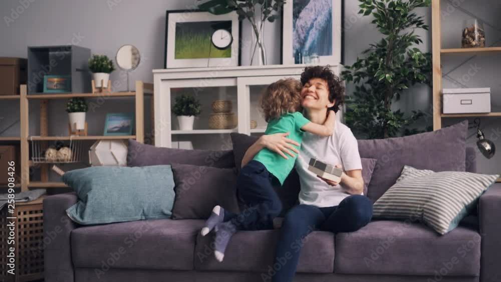 Young woman is using smartphone sitting on sofa when child giving her present kissing and hugging making surprise. Happiness, gifts and family concept. Young woman is using smartphone sitting on sofa when child giving her present kissing and hugging making surprise. Happiness, gifts and family concept.