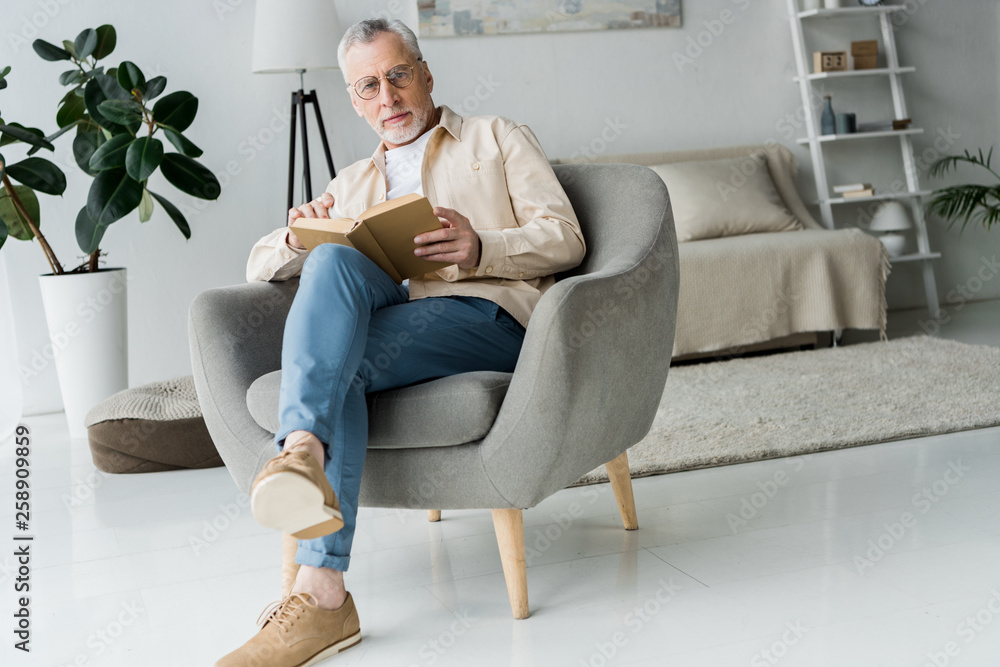 pensive retired man in glasses holding book while sitting in armchair at home pensive retired man in glasses holding book while sitting in armchair at home