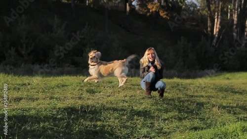 A young woman, blonde, squatting in a clearing next to her Golden Retriever dog, barks at someone then runs up to his mistress
