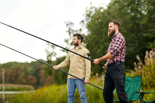 leisure and people concept - happy friends with fishing rods at lake or river