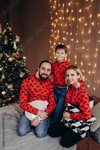 Attractive parents and their little son in red sweaters have fun resting on the bed before a Christmas tree