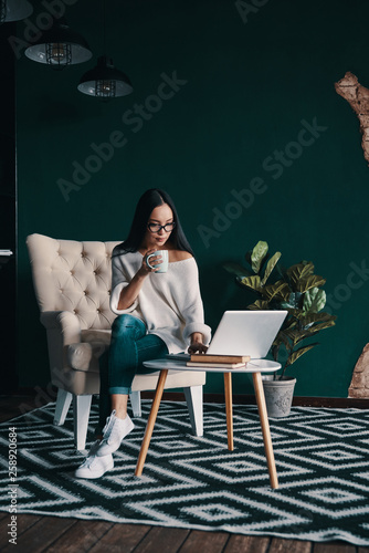 Young and confident. Beautiful young woman using laptop and holding cup while sitting in the armchair