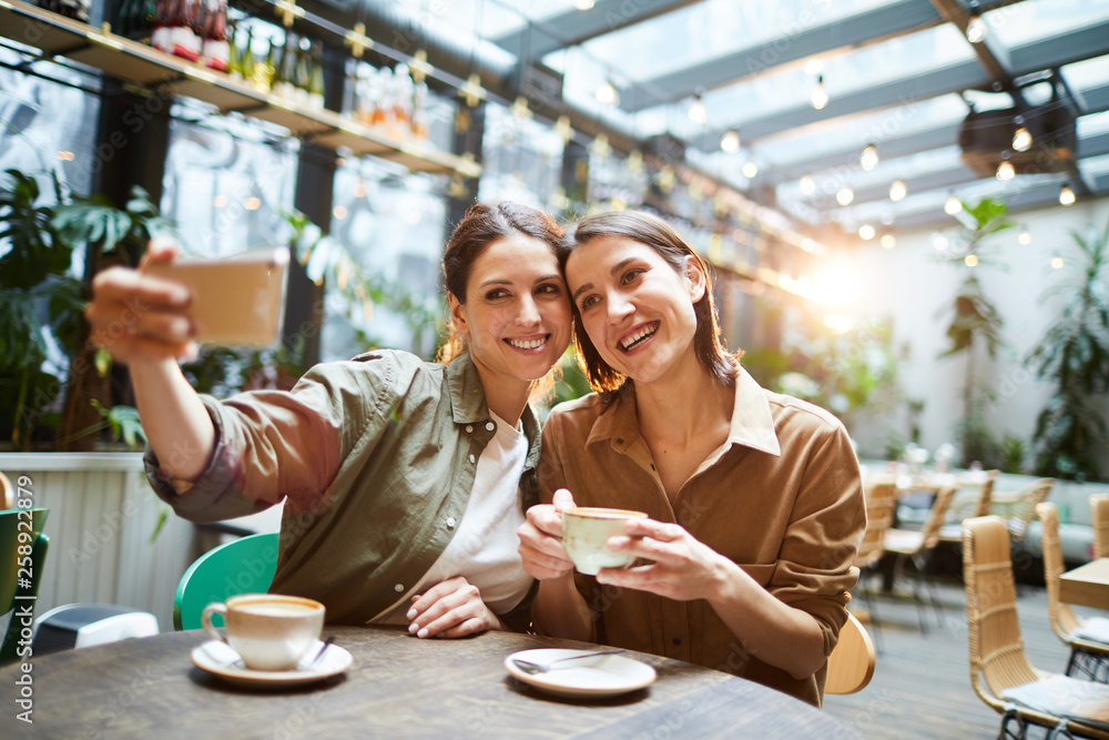 Happy attractive young women in casual shirts sitting at table in coffee shop and photographing together while drinking coffee