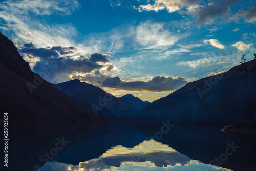 Amazing blue silhouettes of mountains on sunset. Vivid dawn sky reflected in mountain lake. Wonderful colorful highland landscape. Beautiful ripples on lake water on sunrise. Picturesque mountainscape