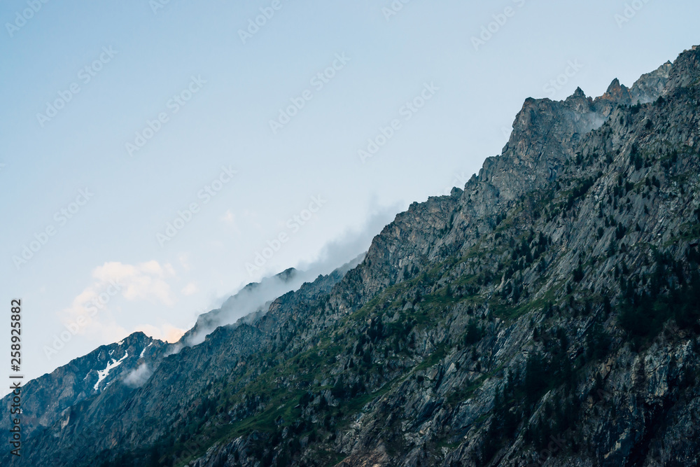 Ghostly giant rocks with trees in thick fog. Mysterious huge mountain with snow in mist. Early morning in mountains. Impenetrable fog. Dark atmospheric eerie landscape. Tranquil mystic atmosphere.