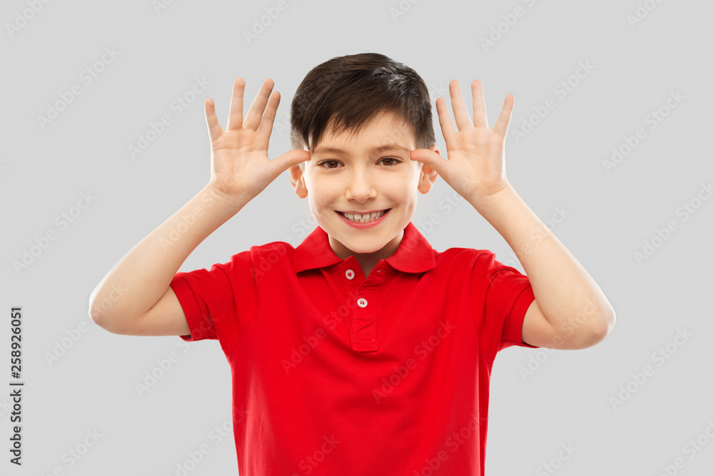 childhood, expressions and fun concept - happy little boy in red polo t-shirt making big ears by hands over grey background childhood, expressions and fun concept - happy little boy in red polo t-shirt making big ears by hands over grey background
