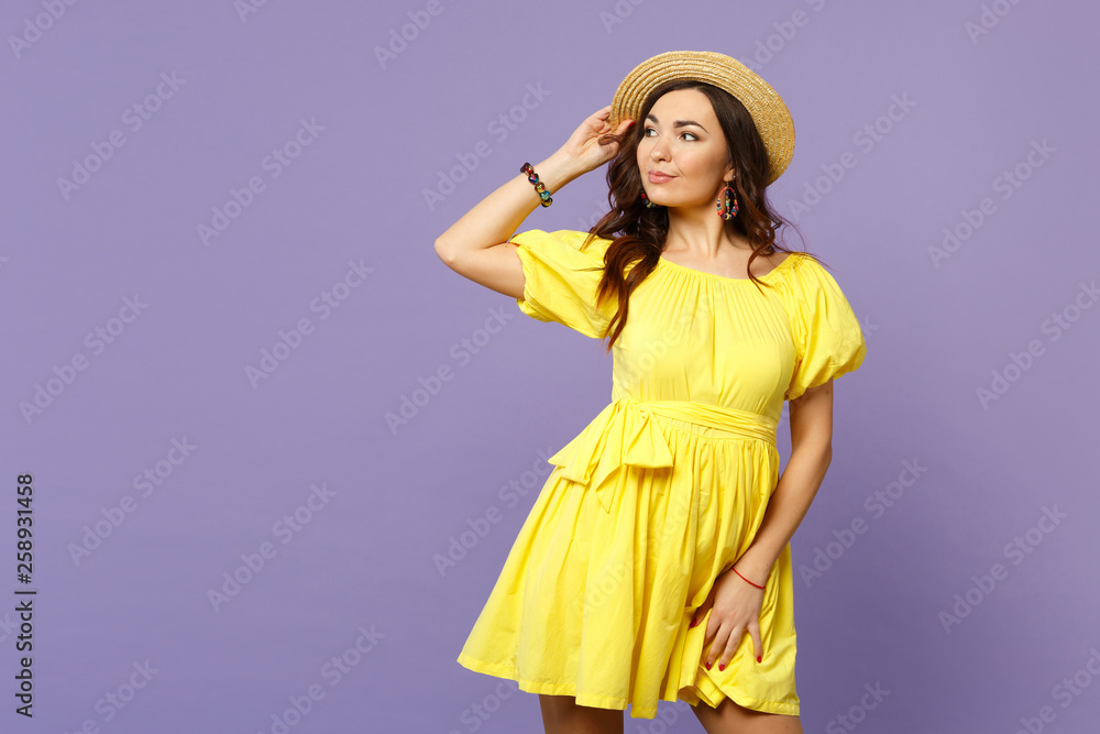 Portrait of pretty young woman in yellow dress keeping hand on summer hat looking aside isolated on pastel violet background in studio. People sincere emotions, lifestyle concept. Mock up copy space. Portrait of pretty young woman in yellow dress keeping hand on summer hat looking aside isolated on pastel violet background in studio. People sincere emotions, lifestyle concept. Mock up copy space.