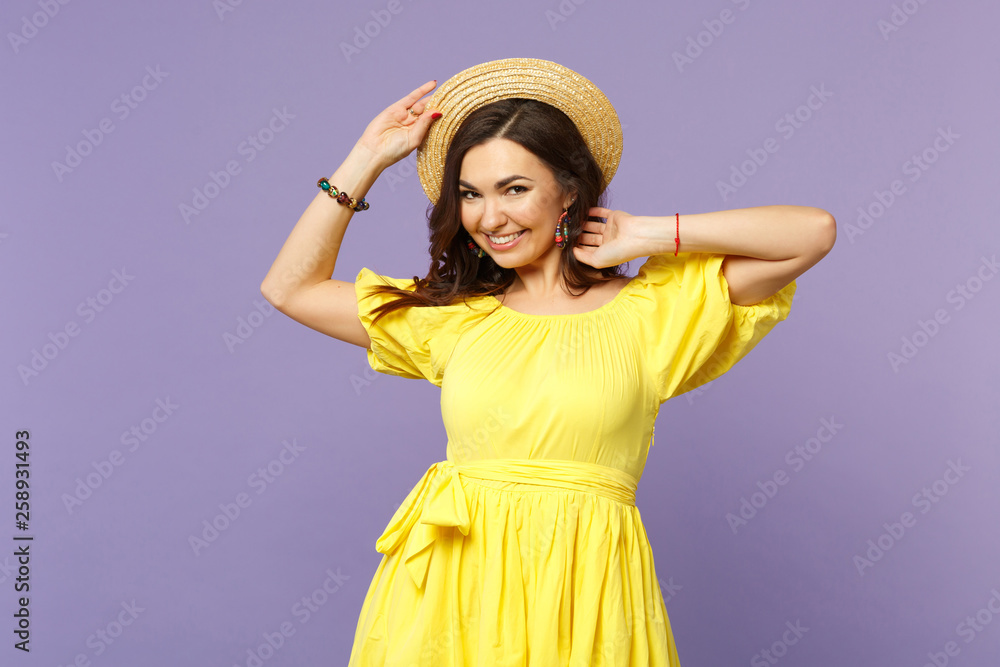 Portrait of smiling young woman in yellow dress keeping hand on summer hat looking camera isolated on pastel violet background in studio. People sincere emotions lifestyle concept. Mock up copy space. Portrait of smiling young woman in yellow dress keeping hand on summer hat looking camera isolated on pastel violet background in studio. People sincere emotions lifestyle concept. Mock up copy space.