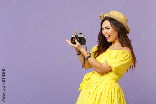 Cheerful young woman in yellow dress hat taking picture on retro vintage photo camera looking aside isolated on pastel violet background. People sincere emotions lifestyle concept. Mock up copy space. photo