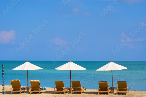 Three white beach umbrellas in a row above lounge chairs on empty sand beach on the ocean shore. Travel and leisure concept. Tropical paradise. Recreation resort.