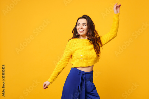 Portrait of smiling young woman in sweater, blue trousers looking aside, rising spreading hands isolated on yellow orange background. People sincere emotions, lifestyle concept. Mock up copy space.