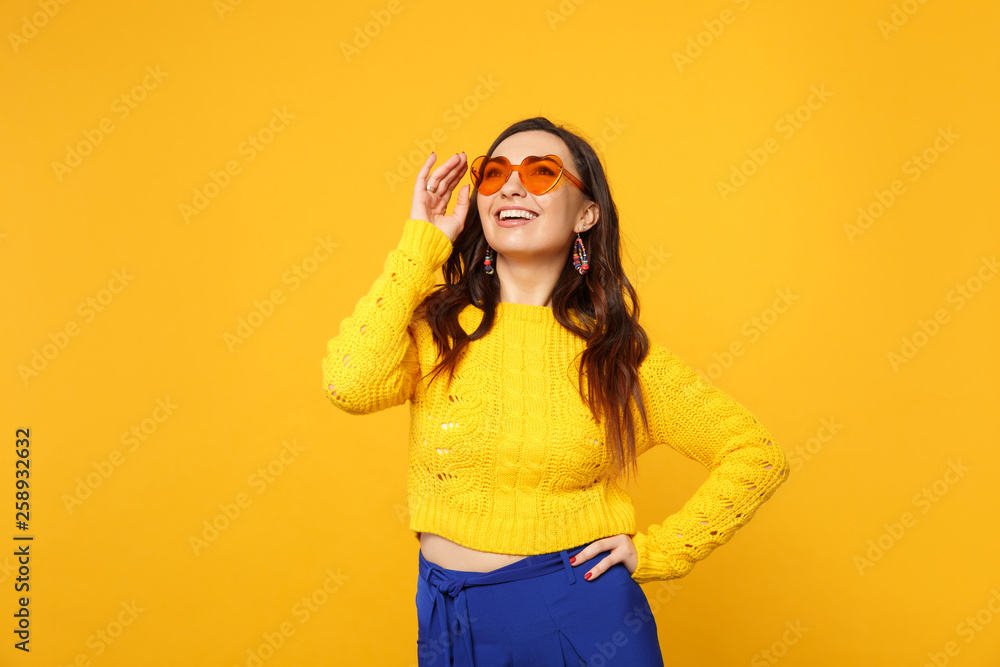 Portrait of joyful young woman in sweater, blue trousers, heart glasses standing, looking up isolated on yellow orange wall background. People sincere emotions, lifestyle concept. Mock up copy space. Portrait of joyful young woman in sweater, blue trousers, heart glasses standing, looking up isolated on yellow orange wall background. People sincere emotions, lifestyle concept. Mock up copy space.