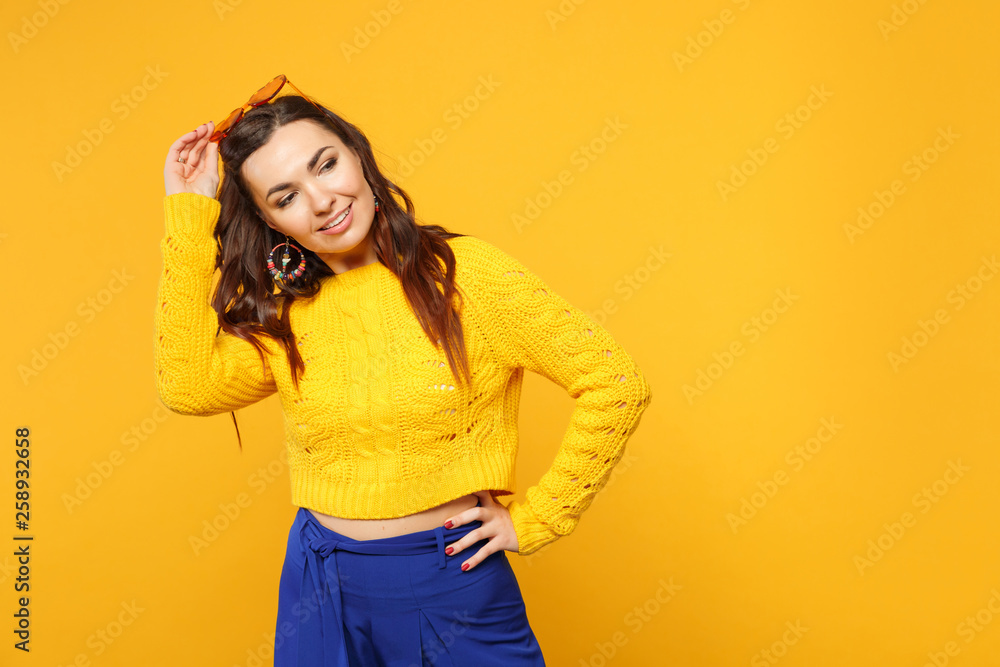 Portrait of smiling young woman in sweater, blue trousers, heart glasses standing, looking aside isolated on yellow orange background. People sincere emotions, lifestyle concept. Mock up copy space. Portrait of smiling young woman in sweater, blue trousers, heart glasses standing, looking aside isolated on yellow orange background. People sincere emotions, lifestyle concept. Mock up copy space.