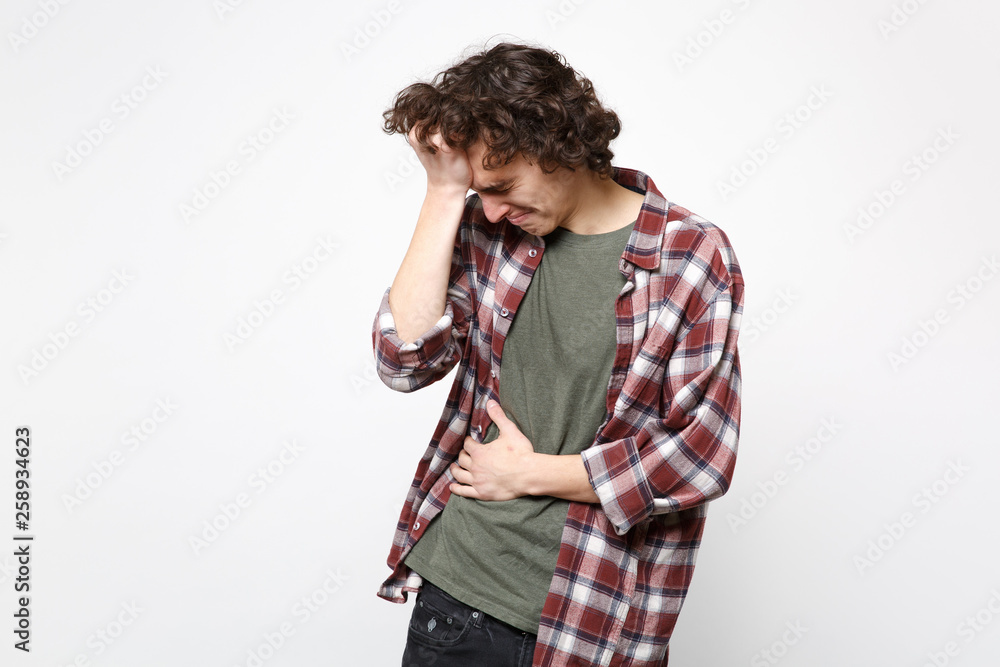 Portrait of crying young man in casual clothes putting hand on head holding hand on stomach isolated on white wall background in studio. People sincere emotions, lifestyle concept. Mock up copy space. Portrait of crying young man in casual clothes putting hand on head holding hand on stomach isolated on white wall background in studio. People sincere emotions, lifestyle concept. Mock up copy space.