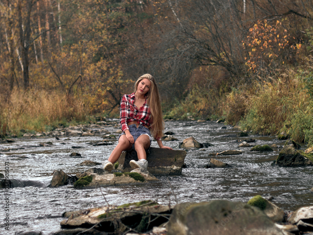 Slim athletic blonde in shorts and a plaid shirt sits in the middle of the river on the stone. Slim athletic blonde in shorts and a plaid shirt sits in the middle of the river on the stone.