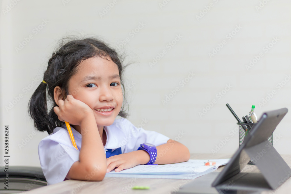 Asian little girl in student uniform doing homework and using tablet on wooden table select focus shallow depth of field Asian little girl in student uniform doing homework and using tablet on wooden table select focus shallow depth of field