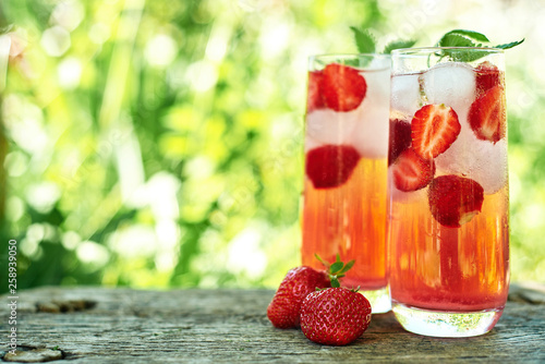 Fresh strawberry lemonade with ice and mint in glasses on wood table with green background outdoors, copy space. Cold summer drink. Sparkling glass with berry cocktail