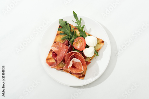 Top view of toast with cherry tomato, arugula and prosciutto on plate on white surface