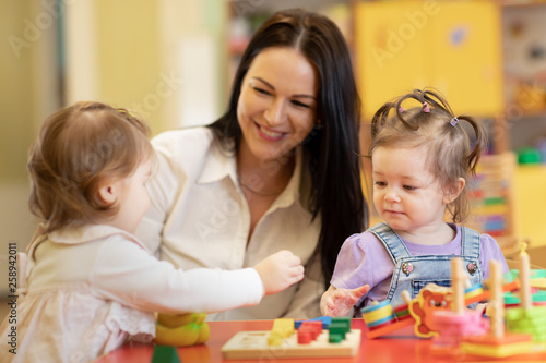 Cute woman and smiling kids playing educational toys at kindergarten or nursery room