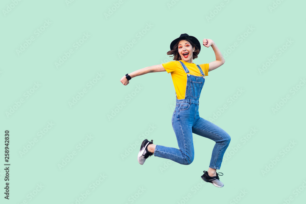 Portrait of happy surprised pretty young hipster girl in blue denim overalls, yellow shirt and black hat jumping and looking at camera and enjoy. indoor studio shot isolated on light green background.