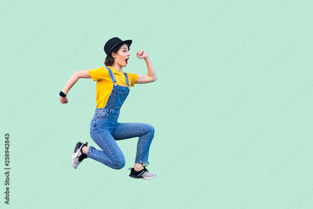 Profile side view portrait of surprised funny young hipster girl in blue denim overalls, yellow shirt and black hat jumping in super mario style. indoor studio shot isolated on light green background.