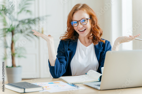 Image of cheerful redhead businesswoman watches webinar or tutorial video, uses free internet connection, raises palms with hesitation, sits at desktop with papers. Female owner of small business