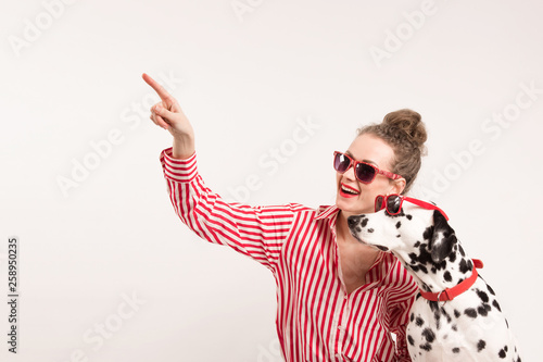 Happy girl and pet in red sunglasses. Young woman hugs dalmatian dog and points with finger on left, indicates copy space on blank wall, isolated over white background. Check this out. Place for text
