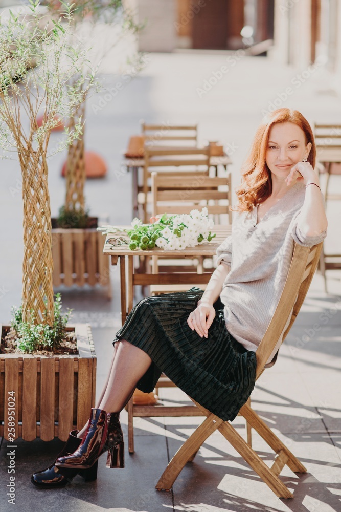 Vertical shot of lovely red haired woman sits on chair waits for