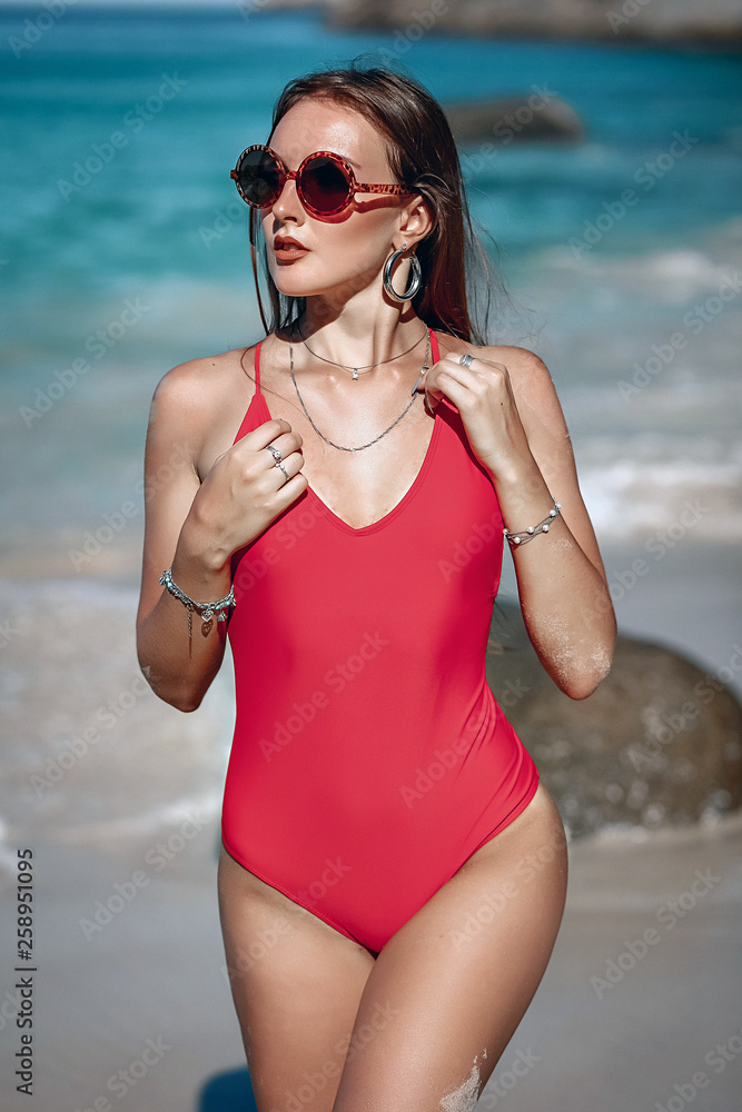 Young girl with a gorgeous body is resting on the beach with white sand near the ocean. Big sea waves. Beautiful sexy model in a red bathing suit and black sunglasses sunbathing. Phuket. Tailand Young girl with a gorgeous body is resting on the beach with white sand near the ocean. Big sea waves. Beautiful sexy model in a red bathing suit and black sunglasses sunbathing. Phuket. Tailand