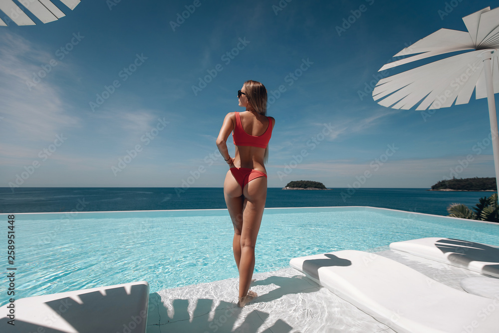 Glamorous, sexy and hot blonde girl with perfect body, in  red swimsuit, posing in the infinity swimming pool. Beautiful seascape. Sexy ass. Trip to warm destination. Phuket. Thailand. Back view. - obrazy, fototapety, plakaty 