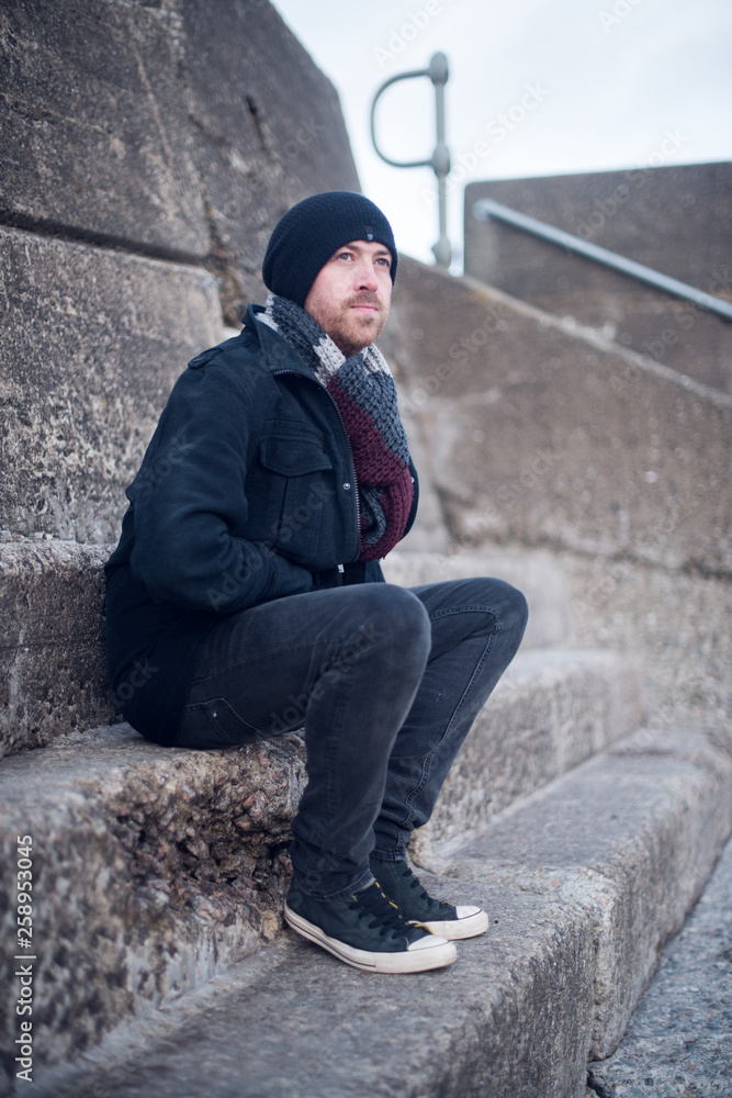 A young handsome man sitting alone and contemplating life by the quiet sea, surrounded by raw concrete sea defence wall. Mental health and depression road to recovery. A young handsome man sitting alone and contemplating life by the quiet sea, surrounded by raw concrete sea defence wall. Mental health and depression road to recovery.