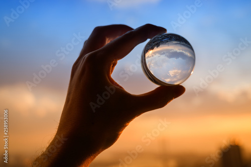 The hand holds glass ball which reflects sunset sky over city.