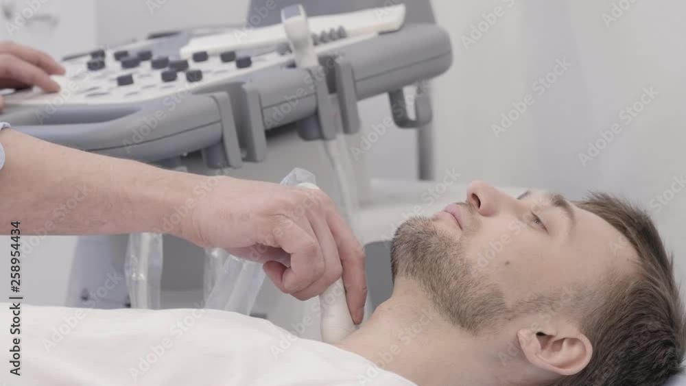 Man lying during undergoing ultrasound diagnostic for thyroid. Ultrasonography doctor using researcher scanner at ratient neck. Female hand holding instrument and carefully checking a problem area Man lying during undergoing ultrasound diagnostic for thyroid. Ultrasonography doctor using researcher scanner at ratient neck. Female hand holding instrument and carefully checking a problem area
