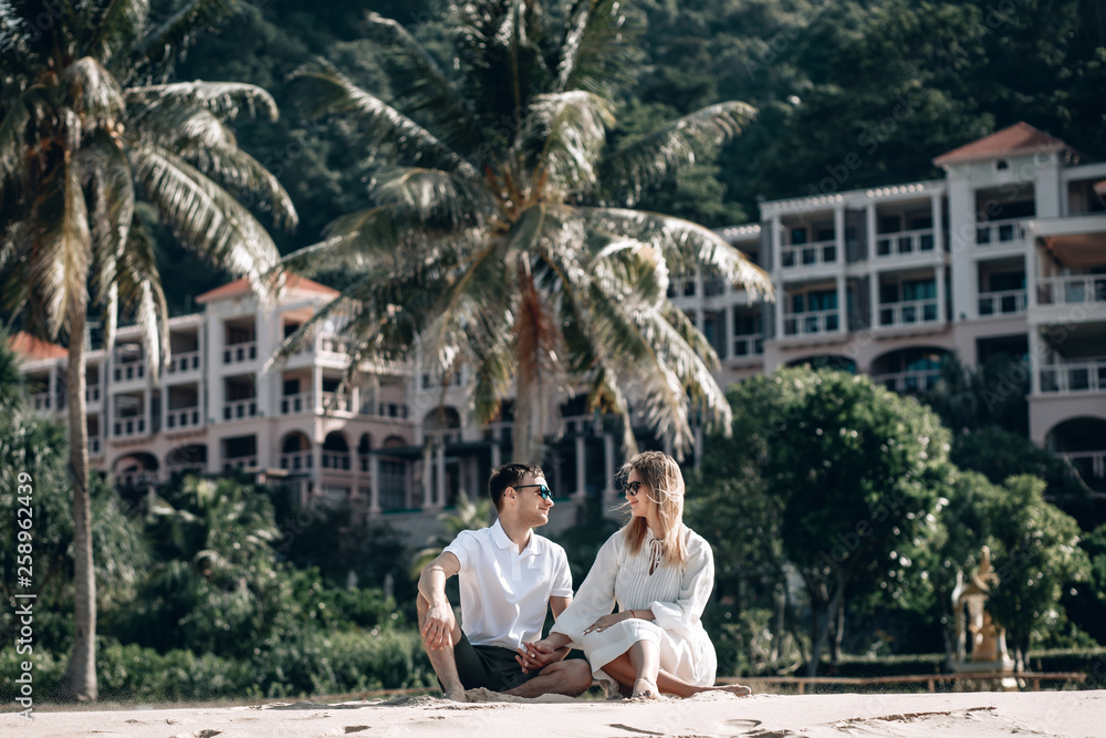 Romantic couple on the beach sitting on the sand. Blurred hotel background. Trip to warm destination