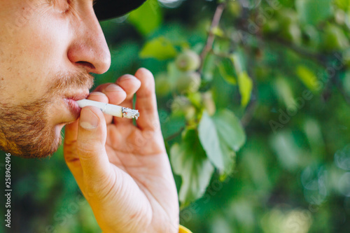 smoldering cigarette in the hand of a man with a beard on nature against the background of green trees. Nicotine tobacco smoke. Unhealthy Lifestyle. Close-up  ash. Smoking break