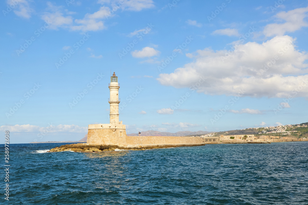 Ancient brick lighthouse. The tourists are standing on the wall. Popular place for holidays seaport Chania, Creete island, Greece. Seafront along the emerald sea with waves. Blue sky with clouds.