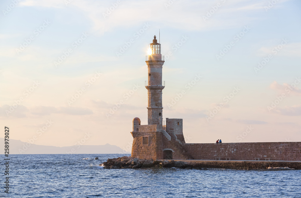 High, beautiful, ancient lighthouse made of bricks. Marvelous sunset lights the sky. Along the sea shore there is a wall and the road. Touristic resort Chania, Creete island, Greece.