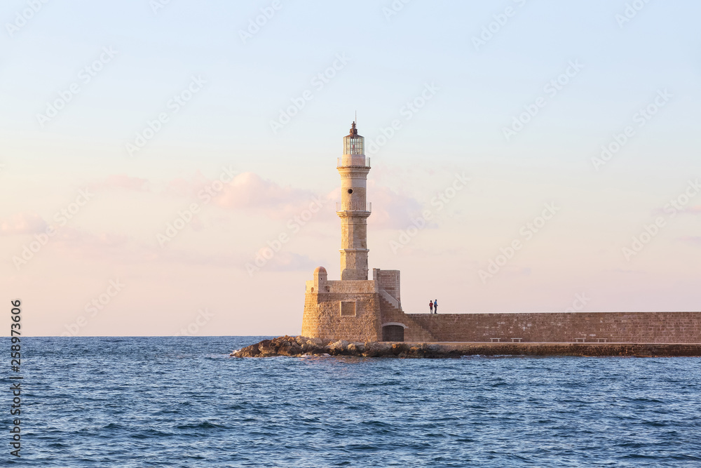 High, beautiful, ancient lighthouse made of bricks. Touristic resort Chania, Creete island, Greece. Marvelous sunset lights the sky. Along the sea shore there is a wall and the road.
