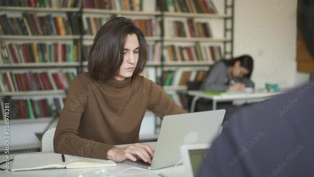 Middle shot of serious young student man typing on laptop. Pan shot of student doing exercises typing on laptop head turning back. Earphones and notebook lying on the table Middle shot of serious young student man typing on laptop. Pan shot of student doing exercises typing on laptop head turning back. Earphones and notebook lying on the table