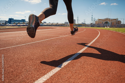 Back view of crop legs of female teen athlete in sports tights running on track at stadium on sunny day