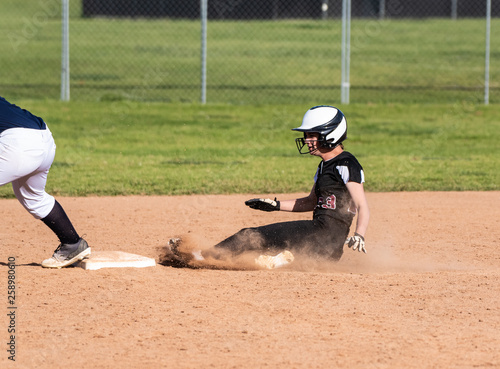 Skilled teenage softball player sliding safely into second base in a cloud of dust.