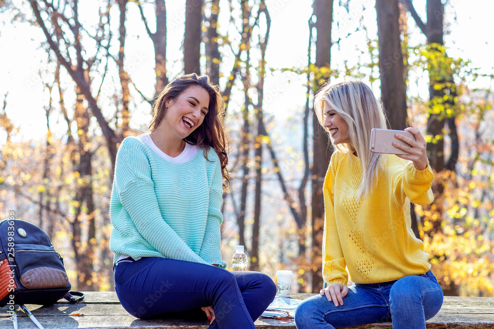Obraz premium Two girls sitting on the desk in forest and looking at mobile phone