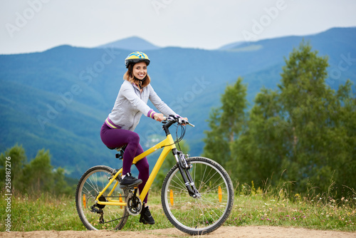 Smiling young female riding on yellow bicycle on a rural trail in the mountains on summer evening, wearing helmet. Mountains, forests on the blurred background. Outdoor sport activity