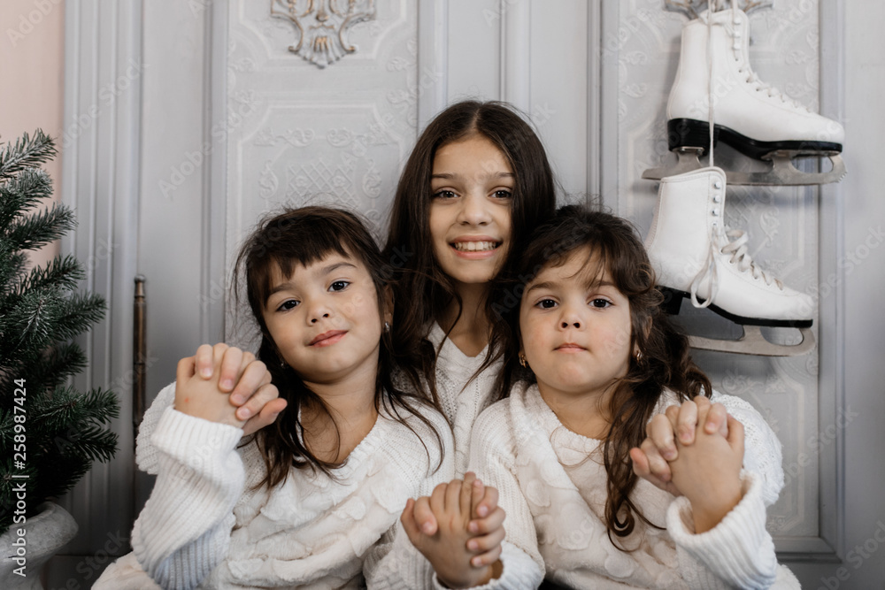 Three charming little sisters in white sweaters and blue jeans sit in a cozy room with Christmas decor Three charming little sisters in white sweaters and blue jeans sit in a cozy room with Christmas decor