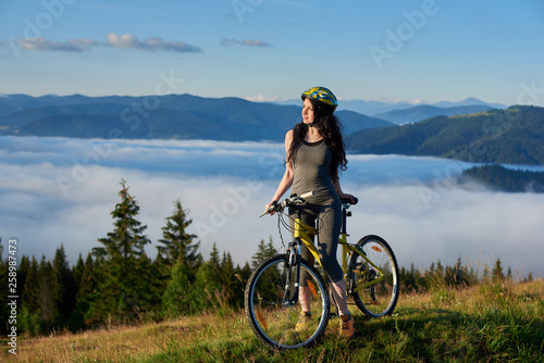 Athlete female cyclist with yellow bicycle in the mountains, wearing helmet, enjoying valley view on sunny morning. Foggy mountains, forests on the blurred background. Outdoor lifestyle activity