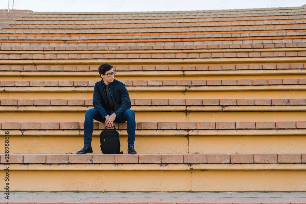 Young businessman with backpack sitting on stairs looking at distance Young businessman with backpack sitting on stairs looking at distance