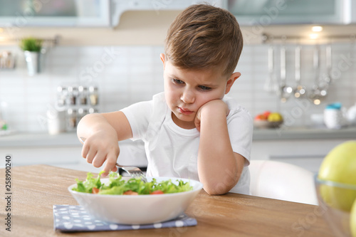 Unhappy little boy eating vegetable salad at table in kitchen