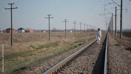Amazing slim unusual beautiful girl in white translucent dress blown by wind and with wax makeup on face in form of bloody wound goes at camera on railroad tracks