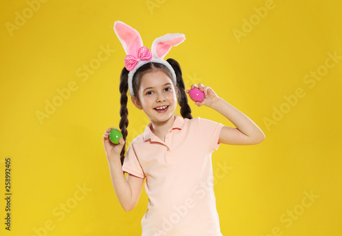 Little girl in bunny ears headband holding Easter eggs on color background