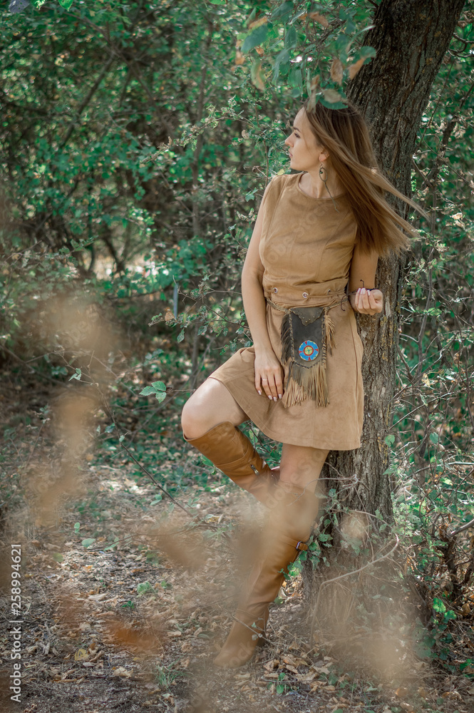 Fototapeta premium A beautiful dark-haired girl in the boho style stands by a tree. Beautiful nature, summer. Ethnic jewelry.